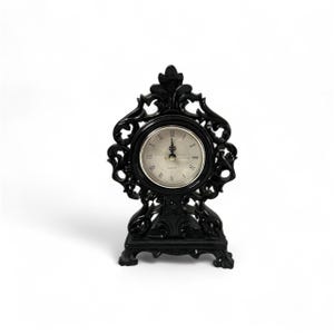 May include: Black decorative table clock with a round face and Roman numeral hour markers. The clock has an ornate, openwork frame and a rectangular base. The clock's hands indicate the time. The clock is made of a dark material.