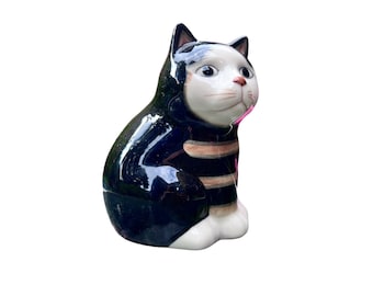 Peut inclure: Figurine de chat en céramique avec un corps noir brillant et un visage, des pattes et une poitrine blancs. Le chat a des rayures marron et beige et un nez rose. Le chat est assis droit et regarde sur le côté.
