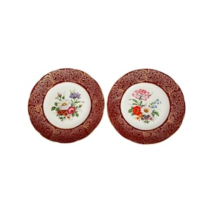 Puede incluir: Dos platos decorativos con diseño floral. Los platos tienen un borde burdeos con un patrón de filigrana dorada. El centro de cada plato presenta un ramo de flores coloridas sobre un fondo blanco. Un diseño clásico y elegante.