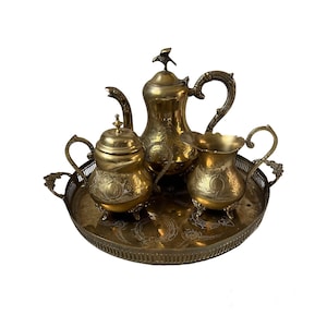 May include: A vintage brass tea set, including a teapot with a bird-shaped finial, a sugar bowl, and a creamer, all on a matching tray. The set features ornate floral and scrollwork detailing, perfect for serving tea or coffee.