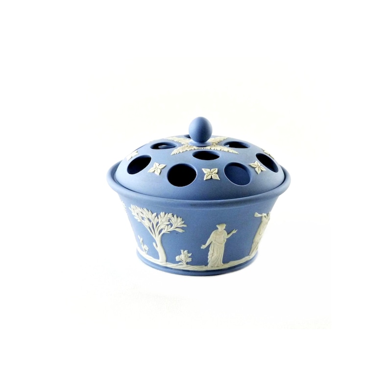May include: A light blue ceramic flower frog with a lid. The lid has several holes and is topped with a small sphere. The sides of the frog feature white relief designs of figures and trees. A decorative piece for floral arrangements.