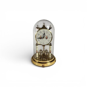 Puede incluir: Un reloj de sobremesa dorado con una cúpula de cristal. La esfera del reloj es blanca con detalles florales y números negros. La base y el péndulo son dorados, con detalles florales decorativos.