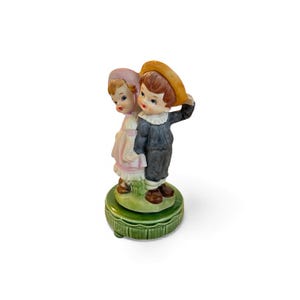 May include: A vintage ceramic figurine of a boy and girl standing back-to-back. The girl wears a pink dress and bonnet, while the boy is in a dark blue suit and holds a yellow hat. The figurine is mounted on a green, circular base.