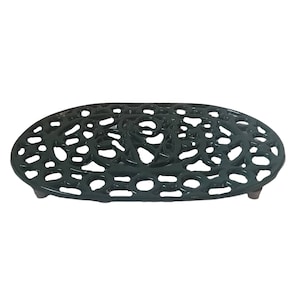 May include: Dark green cast iron trivet. The oval-shaped trivet features an ornate floral and scrollwork pattern. It has small feet and is designed to protect surfaces from hot cookware. A decorative kitchen accessory.