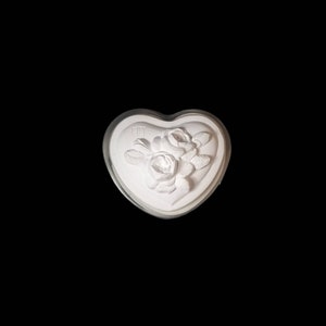 Puede incluir: Una caja de baratijas blanca en forma de corazón con un diseño floral en relieve con rosas y hojas. La caja tiene un borde transparente y redondeado sobre un fondo negro. El diseño floral está centrado en la tapa.