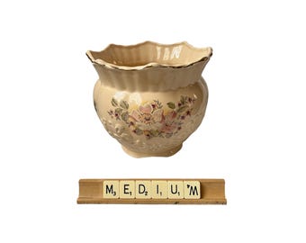 May include: A cream-colored ceramic planter with a scalloped rim and floral design. The planter features a gold trim and embossed details. The word "MEDIUM" is spelled out with Scrabble tiles below the planter.