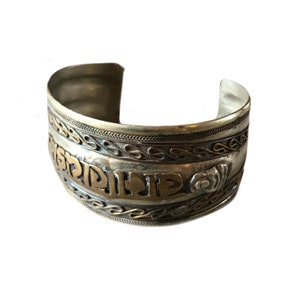 Puede incluir: Un brazalete ancho de plata con letras doradas y adornos decorativos. El brazalete tiene un diseño abierto y presenta patrones intrincados y un acento floral. El metal tiene un aspecto ligeramente deslustrado.