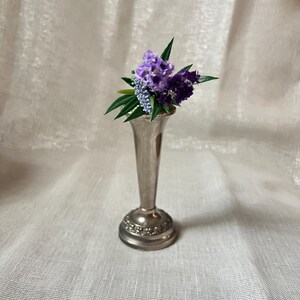 May include: A small, silver-colored bud vase holds a bouquet of purple and blue flowers with green leaves. The vase has a flared base with a decorative pattern and a slender, elongated neck.
