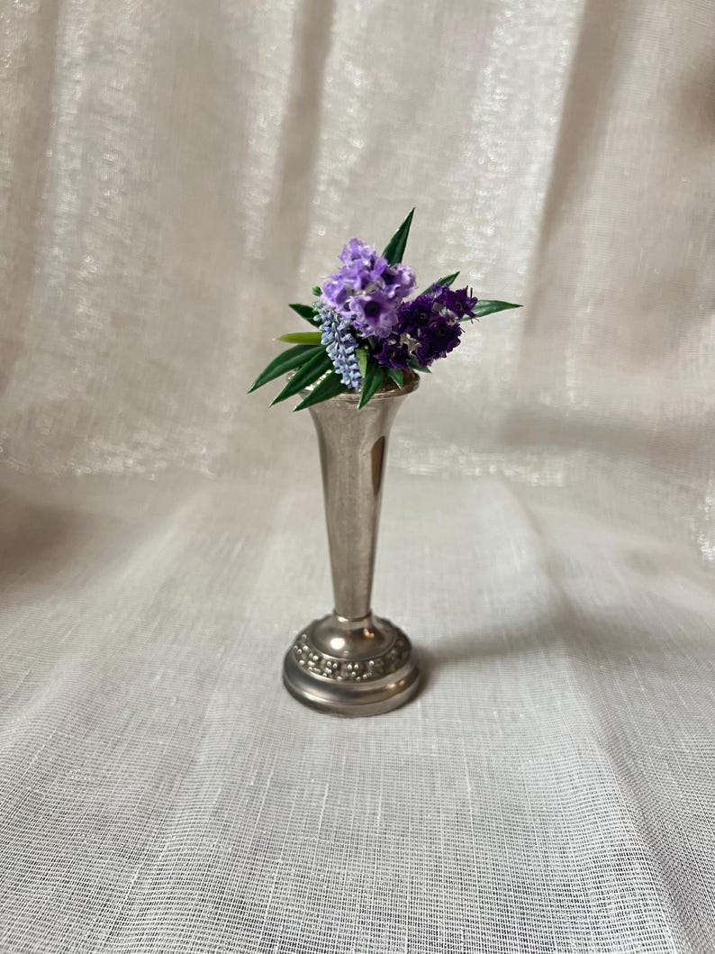 May include: A small, silver-colored bud vase holds a bouquet of purple and blue flowers with green leaves. The vase has a flared base with a decorative pattern and a slender, elongated neck.