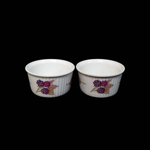 May include: Two white ceramic ramekins with a ribbed design. Each ramekin features a painted design of purple blackberries and leaves. The ramekins are set against a black background.