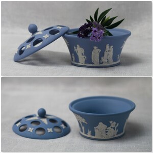 May include: A blue ceramic trinket box with a lid, featuring a classical relief design in white. The lid has decorative cutouts. The box contains purple flowers and green leaves. A decorative piece for the home.