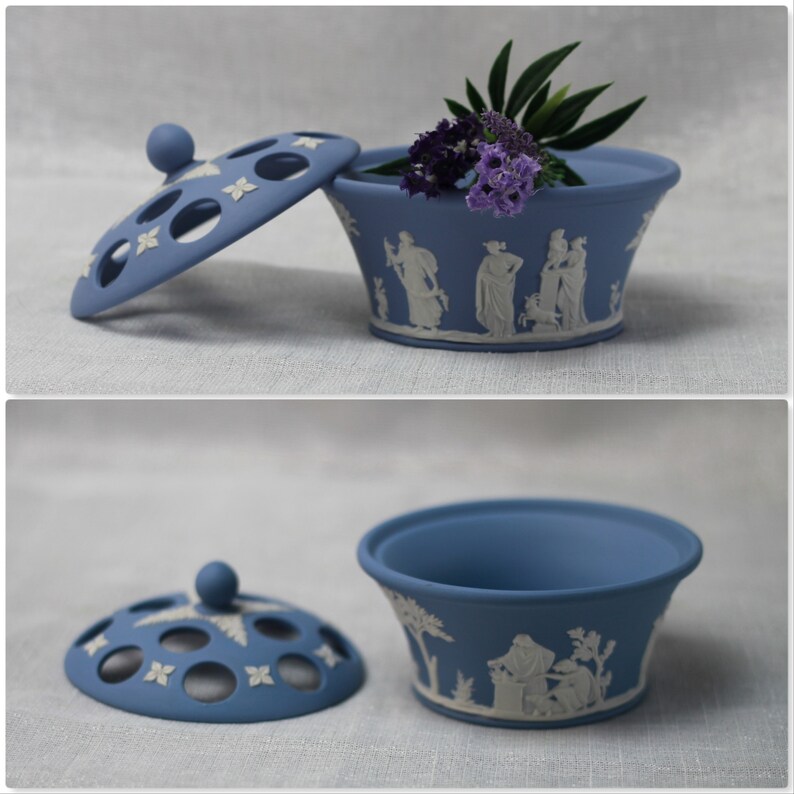 May include: A blue ceramic trinket box with a lid, featuring a classical relief design in white. The lid has decorative cutouts. The box contains purple flowers and green leaves. A decorative piece for the home.