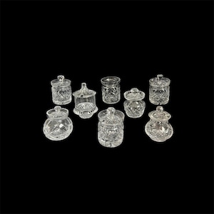 Puede incluir: Una colección de nueve tarros de vidrio transparente con tapas, cada uno con un diseño único. Los tarros varían en forma y tamaño, con patrones y texturas intrincados. Los tarros están dispuestos sobre un fondo negro.