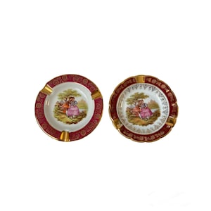 May include: Two vintage ashtrays with a burgundy and gold rim. Each ashtray features a central image of a couple in a garden setting. The ashtrays have gold accents and are likely made of porcelain.