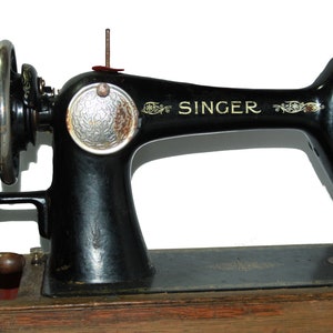 May include: An antique Singer sewing machine, black with gold accents, sits on a wooden base. The machine features a hand wheel, a needle, and the Singer logo. The machine is a classic example of vintage sewing equipment.