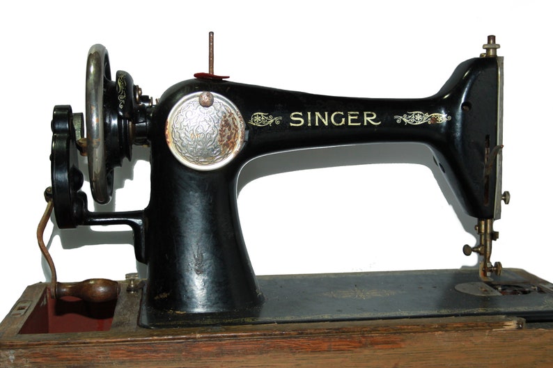 May include: An antique Singer sewing machine, black with gold accents, sits on a wooden base. The machine features a hand wheel, a needle, and the Singer logo. The machine is a classic example of vintage sewing equipment.