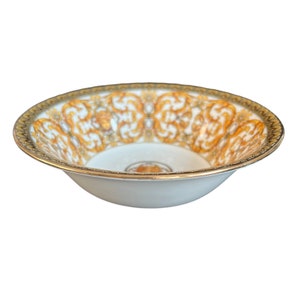 Könnte beinhalten: Weißer Keramikschüssel mit goldenem Rand und aufwendigem gold-weißem Innendesign. Die Schüssel hat ein sich wiederholendes Muster aus goldenen Schnörkeln und ein goldenes Emblem in der Mitte. Ein dekoratives Stück.