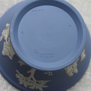 May include: A blue ceramic bowl with white relief designs. The underside features the text "WEDGWOOD" and "MADE IN ENGLAND". The bowl has a circular base and a flared rim, showcasing a classic design.