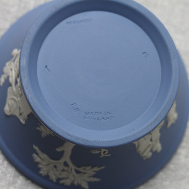 May include: A blue ceramic bowl with white relief designs. The underside features the text "WEDGWOOD" and "MADE IN ENGLAND". The bowl has a circular base and a flared rim, showcasing a classic design.