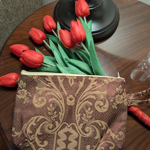 May include: A small, rectangular, brown and gold patterned pouch with a zipper, holding several red tulips with green leaves. The tulips are arranged on a dark wooden surface, near a black lamp base.