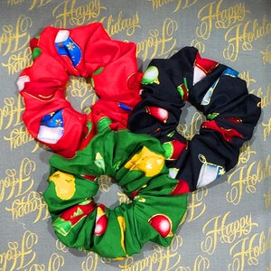 May include: Three festive scrunchies with Christmas patterns. One is red with Santa Claus and gold details, another is green with baubles, and the third is black with gift boxes and decorations. They are displayed on a background with "Happy Holidays" written on it.