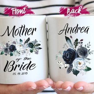 May include: Two white ceramic mugs with blue floral designs. The front mug says "Mother of the Bride" with the date "05.19.2019". The back mug says "Andrea".