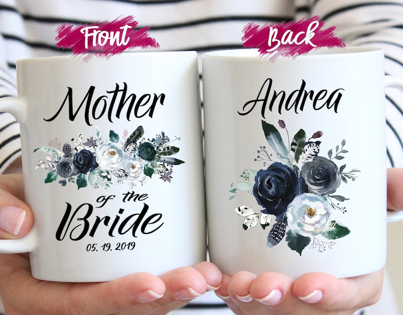 May include: Two white ceramic mugs with blue floral designs. The front mug says "Mother of the Bride" with the date "05.19.2019". The back mug says "Andrea".