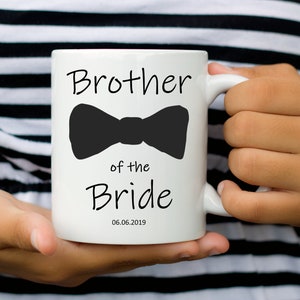 Pu&ograve; includere: Tazza in ceramica bianca con la scritta "Brother of the Bride" e un fiocco nero. La data "06.06.2019" &egrave; stampata sotto. La tazza &egrave; tenuta da una persona che indossa un top a righe bianche e nere.