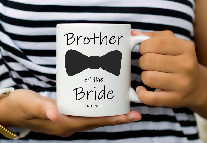 Pu&ograve; includere: Tazza in ceramica bianca con la scritta "Brother of the Bride" e un fiocco nero. La data "06.06.2019" &egrave; stampata sotto. La tazza &egrave; tenuta da una persona che indossa un top a righe bianche e nere.