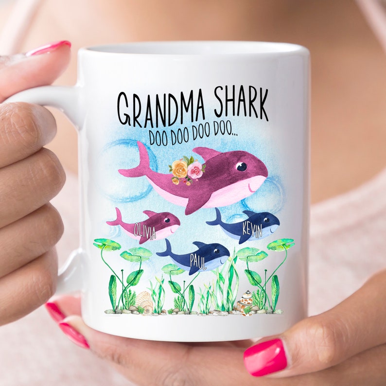 May include: White ceramic mug with a shark family design. The mug features the words "Grandma Shark" and "Doo Doo Doo Doo..." above a purple shark with floral accents, and three smaller sharks with names. The design includes aquatic plants and is a fun gift.