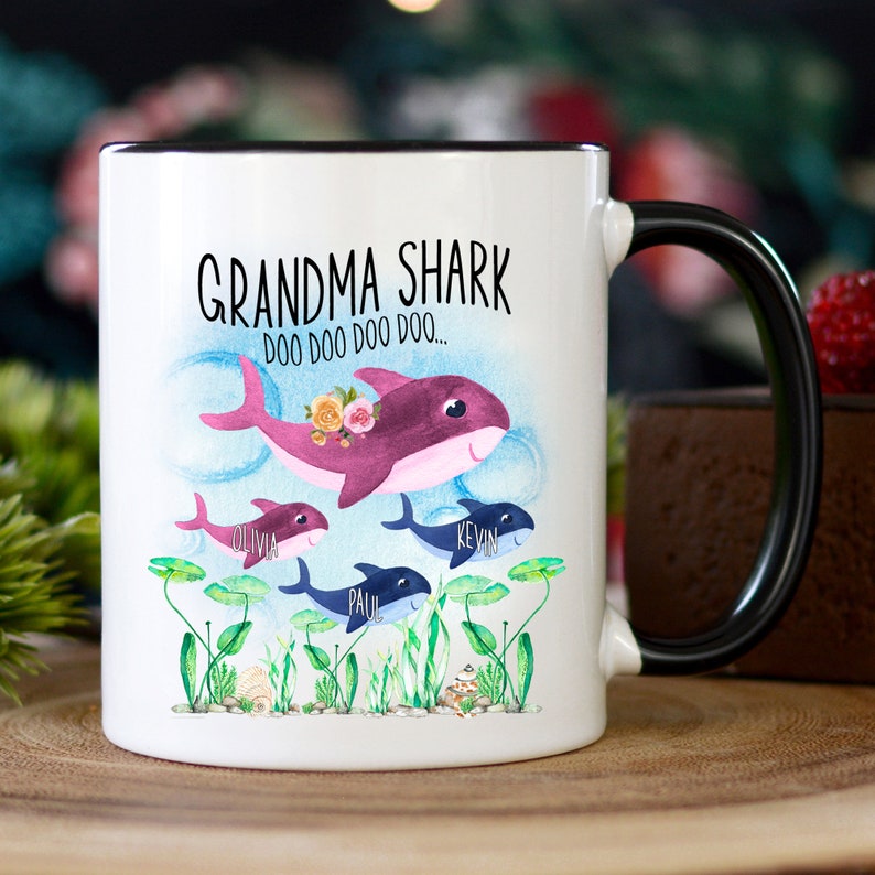 May include: White ceramic mug with a black handle and rim, featuring a whimsical "Grandma Shark" design. The design includes a purple shark with floral accents and smaller shark illustrations with names. The mug is decorated with aquatic plants and the text "DOO DOO DOO DOO..."