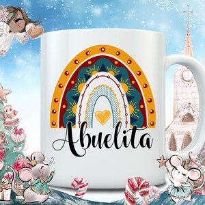 May include: White ceramic mug featuring a colorful rainbow design with a heart and the word "Abuelita" in black script. The rainbow includes shades of yellow, teal, and red. The mug has a standard handle and is likely for hot beverages.