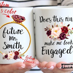 May include: Two white coffee mugs with a floral design. The mug on the left says "future mrs. Smith" with a gold ring and a red gemstone. The mug on the right says "Does this ring make me look engaged?" with a floral design.