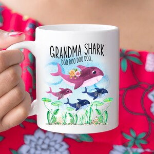 May include: White ceramic mug with a "Grandma Shark" design, featuring a family of cartoon sharks. The top shark has a floral crown. Names are printed below the sharks. The mug has a curved handle.
