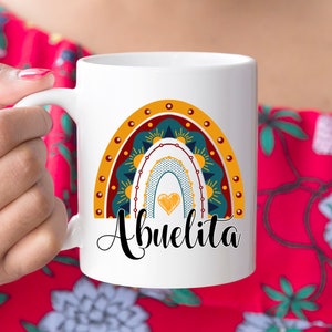 May include: White ceramic mug featuring a colorful rainbow design with the word "Abuelita" in black script. The rainbow includes shades of yellow, teal, and orange, with a heart in the center. The mug has a curved handle.