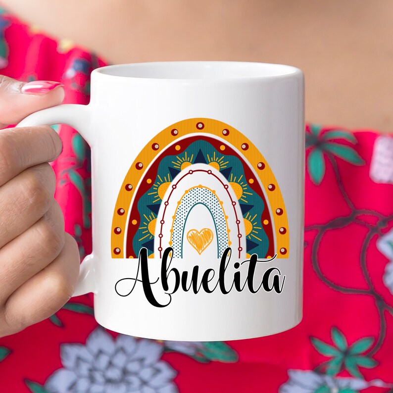 May include: White ceramic mug featuring a colorful rainbow design with the word "Abuelita" in black script. The rainbow includes shades of yellow, teal, and orange, with a heart in the center. The mug has a curved handle.