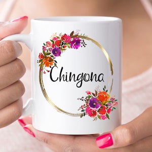 May include: White ceramic mug with a gold circle and floral design. The text "Chingona" is written in black inside the circle.