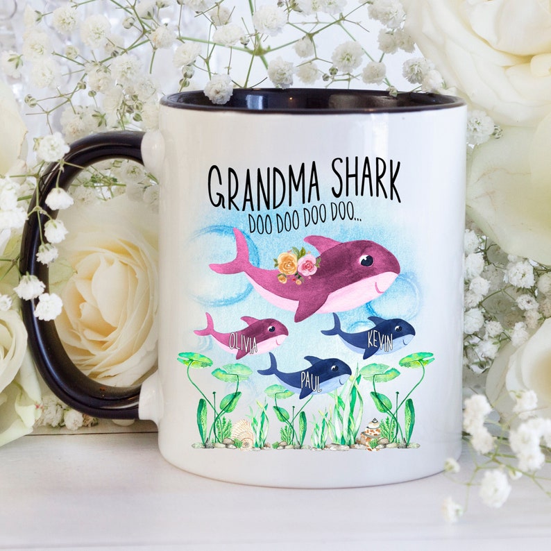 May include: White ceramic mug with a black interior and handle. The mug features a whimsical design with a purple shark labeled "Grandma Shark" and smaller sharks with names. The text "Doo Doo Doo Doo..." is also included.