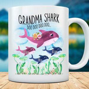 May include: White ceramic mug with a shark family illustration. The design features a purple Grandma Shark with flowers, and three smaller sharks. The mug has the text "Grandma Shark Doo Doo Doo..." and names on the smaller sharks.