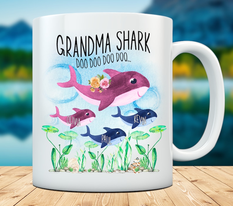 May include: White ceramic mug with a shark family illustration. The design features a purple Grandma Shark with flowers, and three smaller sharks. The mug has the text "Grandma Shark Doo Doo Doo..." and names on the smaller sharks.