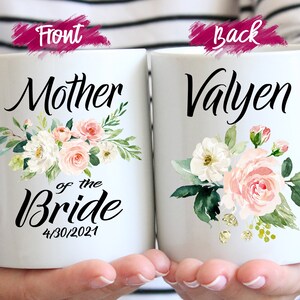 May include: Two white ceramic mugs with floral designs. The front mug says "Mother of the Bride" with the date "4/30/2021". The back mug says "Valyen".