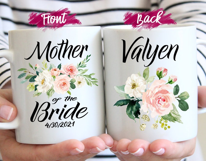 May include: Two white ceramic mugs with floral designs. The front mug says "Mother of the Bride" with the date "4/30/2021". The back mug says "Valyen".