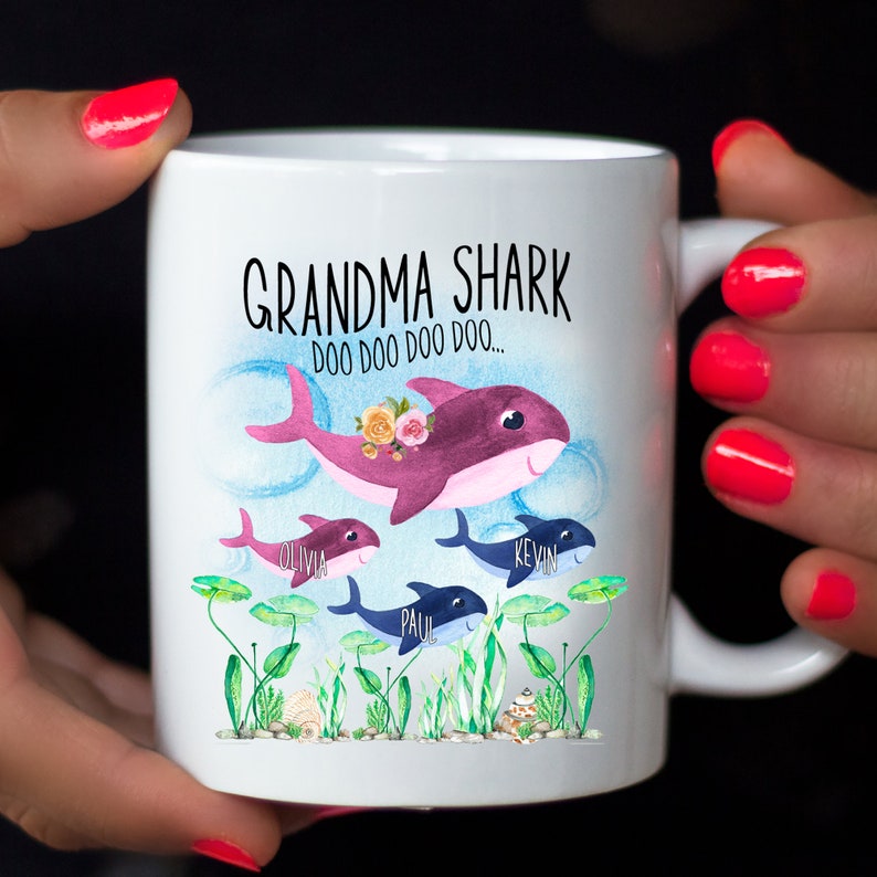 May include: White ceramic mug with a shark family design. The mug features the text "Grandma Shark" and cartoon sharks with names. The largest shark has a floral crown. The mug has a handle on the right side.