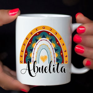 May include: White ceramic mug featuring a colorful rainbow design with a heart and the word "Abuelita" in black script. The rainbow includes shades of teal, yellow, and red. The mug has a handle and is held by hands with red nail polish.