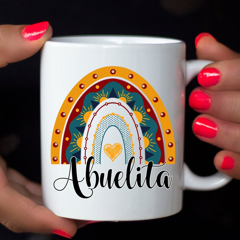 May include: White ceramic mug featuring a colorful rainbow design with a heart and the word "Abuelita" in black script. The rainbow includes shades of teal, yellow, and red. The mug has a handle and is held by hands with red nail polish.