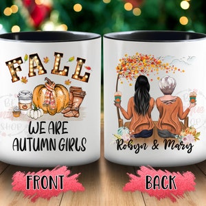 May include: White ceramic mug with a black interior and handle. The front features the word "FALL" in illuminated letters, a pumpkin, and the text "WE ARE AUTUMN GIRLS." The back shows two women with coffee, the names "Robyn & Mary", and fall foliage.