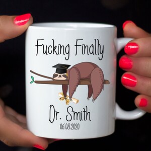 May include: White ceramic mug featuring a cartoon sloth wearing a graduation cap and holding a diploma. The mug has the text "Fucking Finally" above the sloth, with "Dr. Smith" and the date 06.08.2020 below.