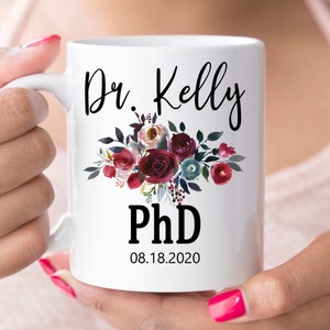 May include: White ceramic mug featuring the name "Dr. Kelly" and "PhD" in black script. A floral arrangement in burgundy, pink, and teal adorns the mug. The date "08.18.2020" is printed below the PhD.