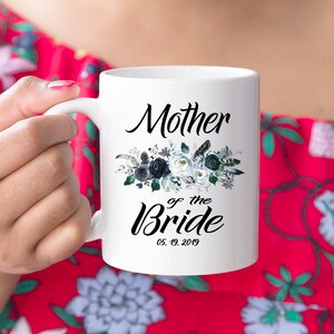 May include: White ceramic mug with a black floral design and the text "Mother of the Bride" with the date "05.19.2019".