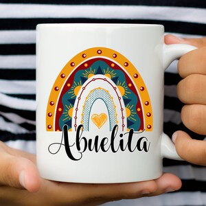 May include: White ceramic mug featuring a colorful rainbow design with a heart and the word "Abuelita" in black script. The rainbow includes shades of teal, orange, and yellow. The mug is held by a person.